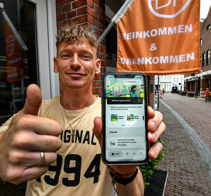 smaboo Kunde social scheduling plattformMaximilian Brockmann hält den Daumen in die Höhe und in der anderen Hand ein Smartphone mit der smaboo App