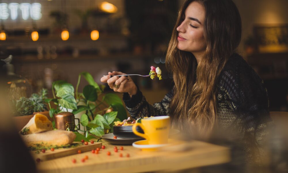 Eine junge Frau isst genießerisch in einem Restaurant ein Nudelgericht, auf dem Tisch vor ihr stehen Pflanzen, Speisen und eine gelbe Tasse.